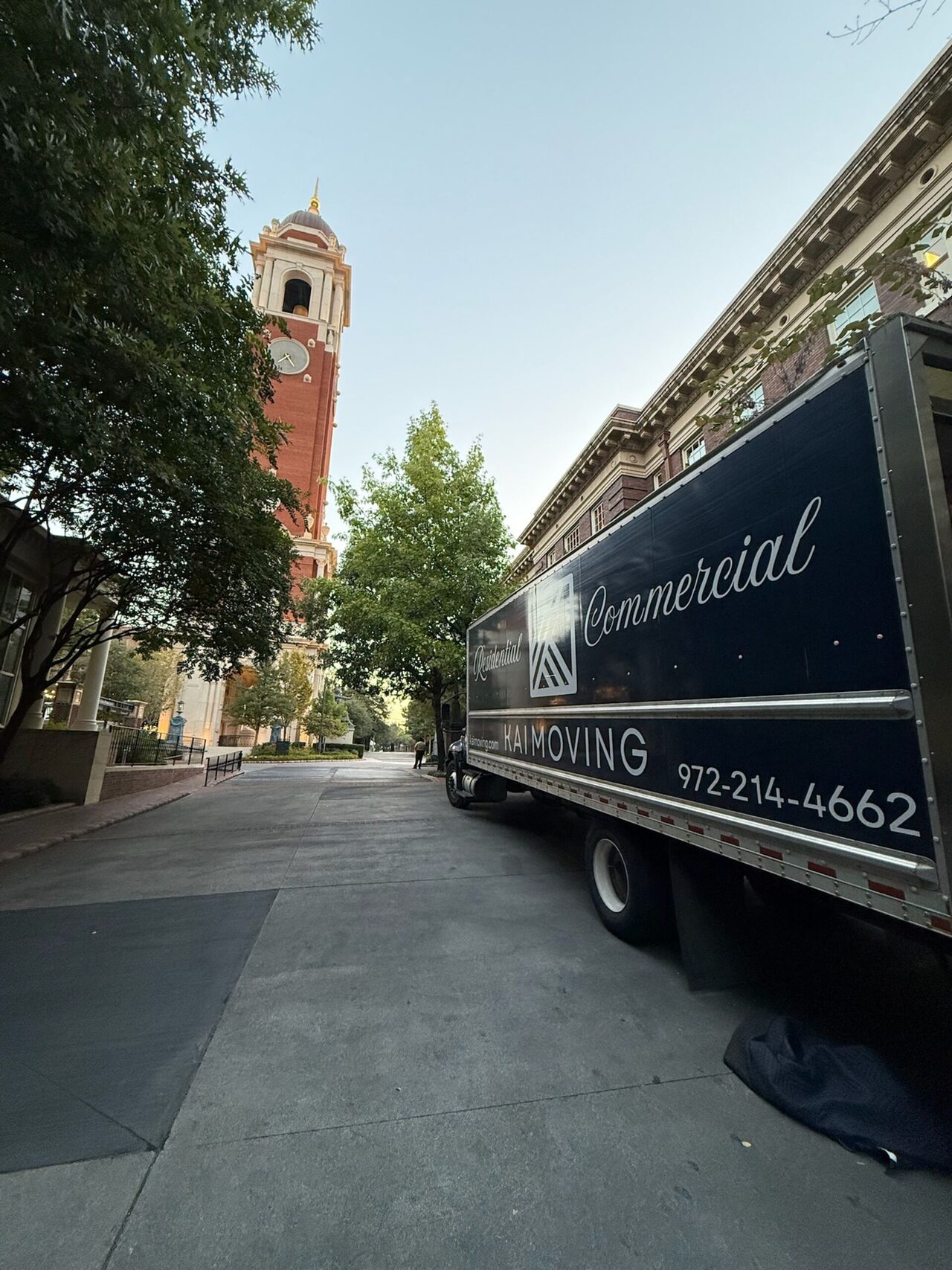 A Kai Moving commercial box truck parked beside a historic Dallas building at dusk.